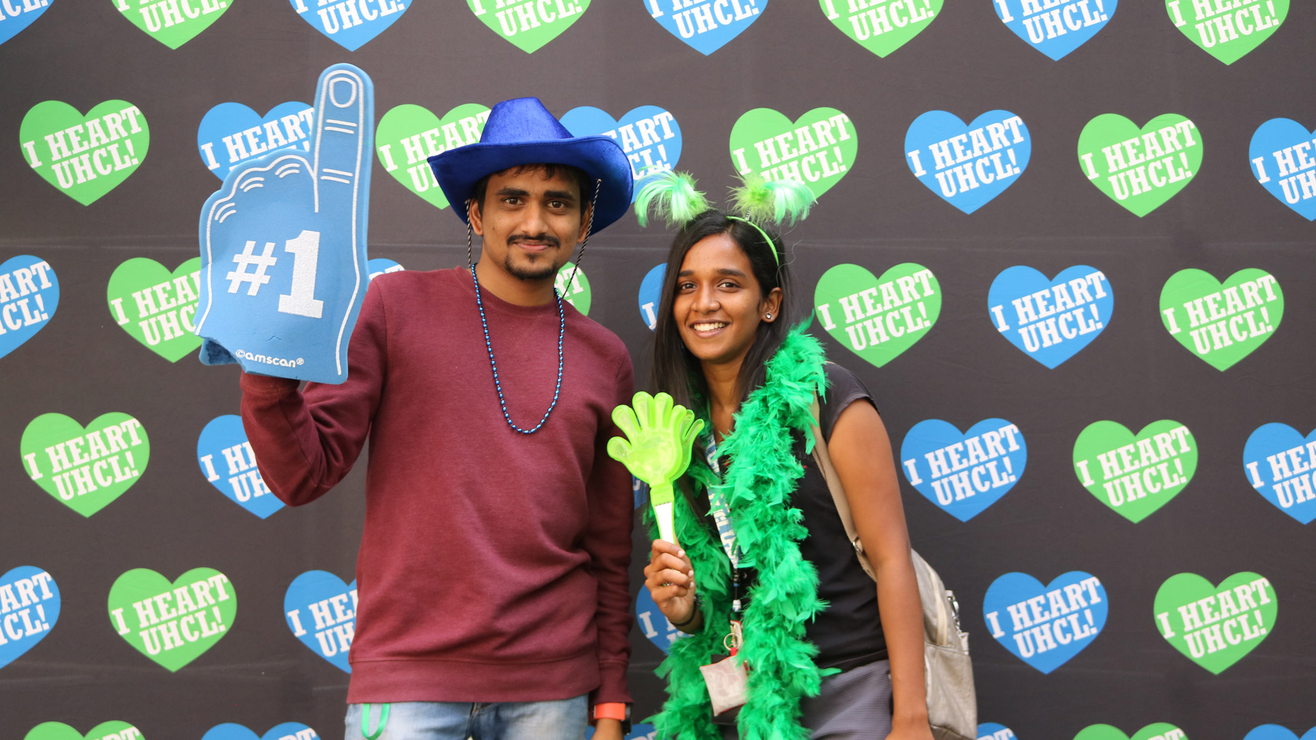 Two students sport blue and green hats, boas, and foam fingers for I Heart UHCL Day